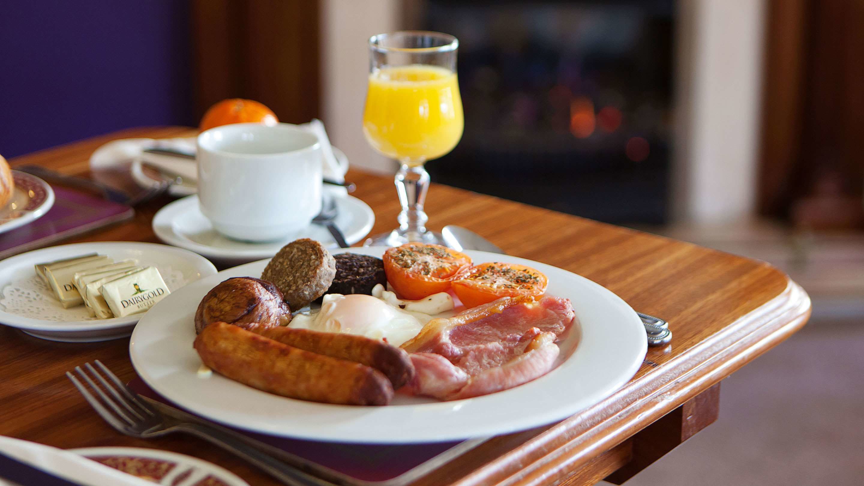 A breakfast plate with eggs, bacon, sausages, and tomatoes, accompanied by a glass of orange juice and a cup of coffee on a wooden table.