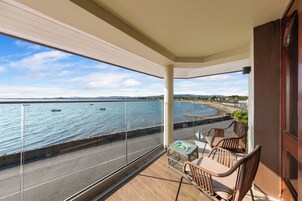A balcony with a view of the sea, a table with chairs, and a small table with items on it.