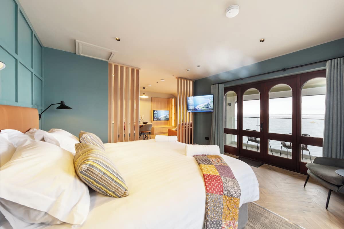 Une chambre d’hôtel avec un grand lit, une télévision, un bureau avec une chaise et une vue sur la mer, visible à travers une porte vitrée.