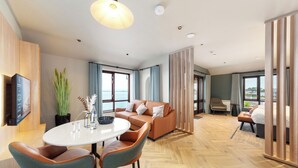 Una habitación de hotel moderna con suelo de madera, una mesa redonda con sillas, un sofá, un televisor montado en la pared y grandes ventanales con vistas al mar.