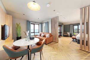 A modern hotel room with a wooden floor, a round dining table with chairs, a sofa, a TV mounted on the wall, and large windows offering a view of the sea.