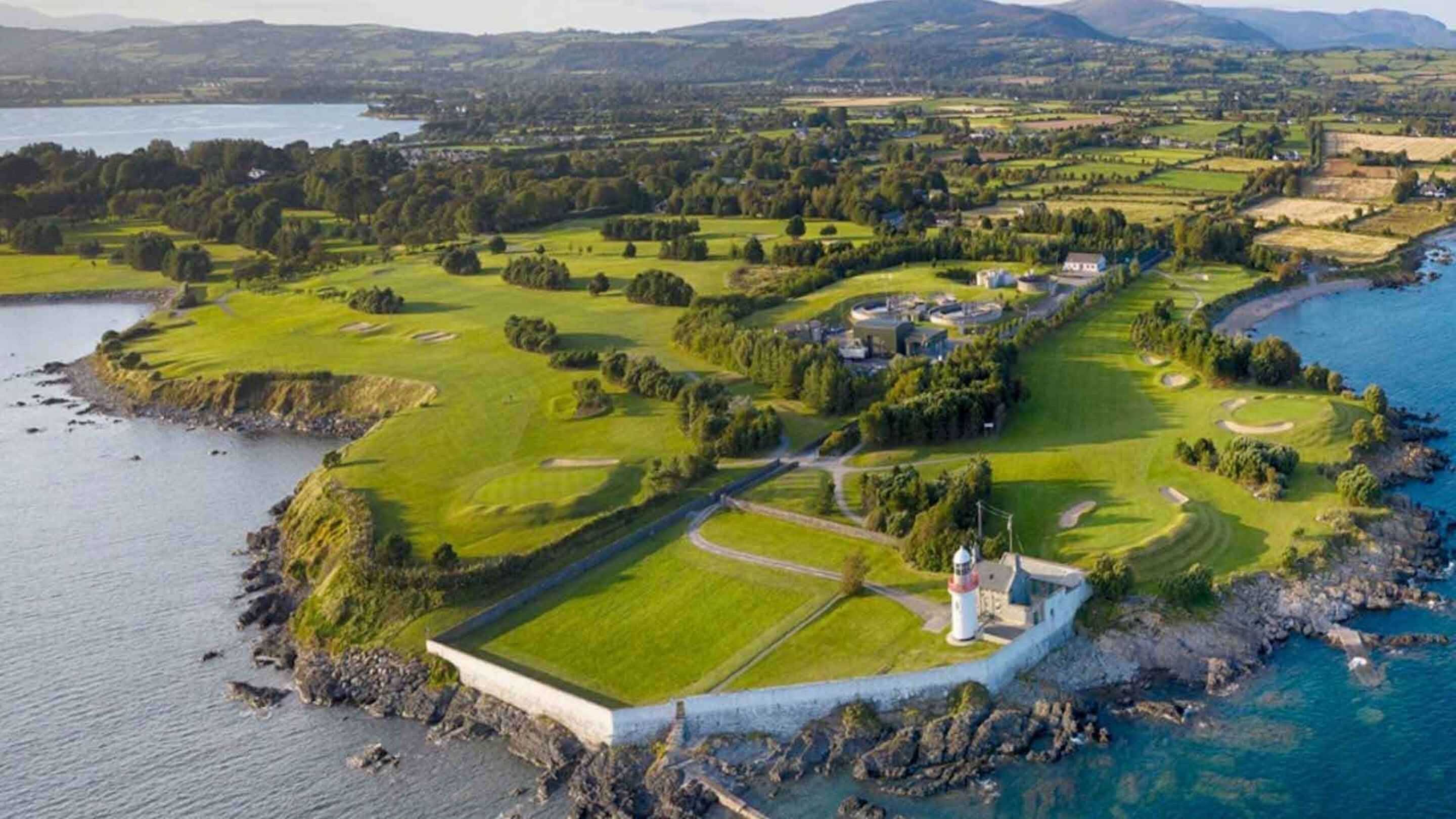 Oversigtsbillede af en kystgolfbane med et fyrtårn, omgivet af frodig vegetation og et vandareal.