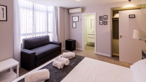 Habitación de hotel con cama, sofá, una mesita, un altavoz y un baño visible a través de una puerta abierta.