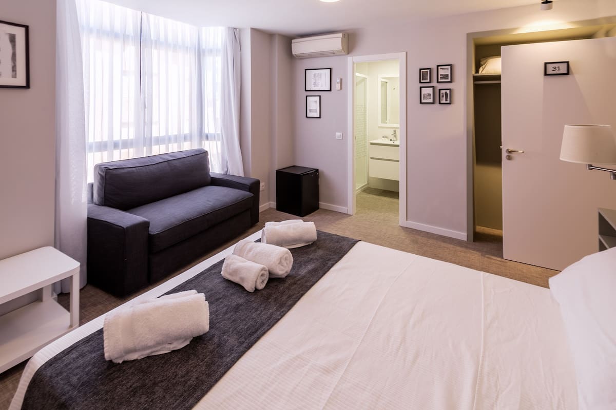 A hotel room with a bed, a sofa, a small table, a speaker, and a bathroom visible through an open door.