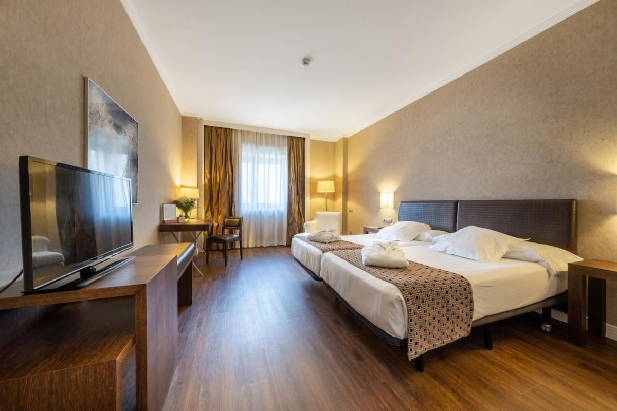 A hotel room with a bed, a desk with a flat-screen TV, a seating area with a chair, and a lamp.