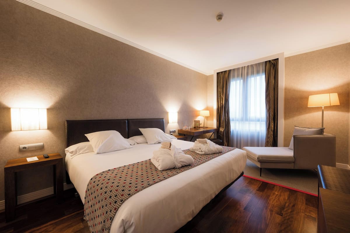 Una habitación de hotel con una cama grande, una mesita de noche, una silla y un escritorio con lámpara.