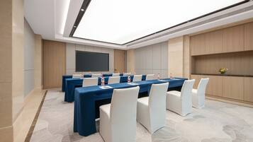 Un salón de conferencias con una larga mesa, sillas blancas y una pantalla grande.