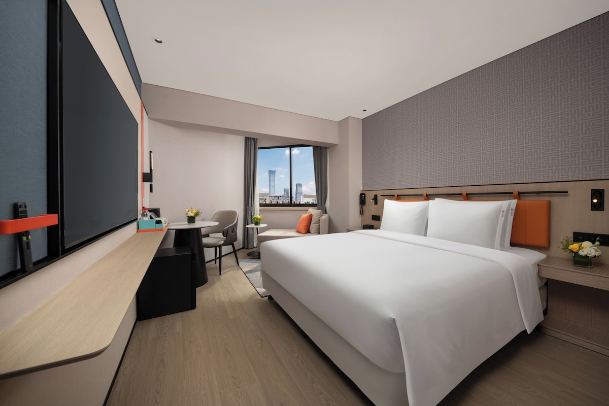 A modern hotel room with a large bed, a desk, a chair, and a view of the cityscape.