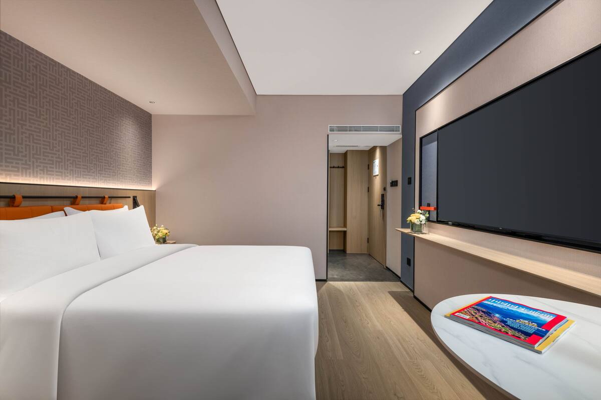 A modern hotel room with a large bed, a flat-screen TV mounted on the wall, and a small table with a magazine.