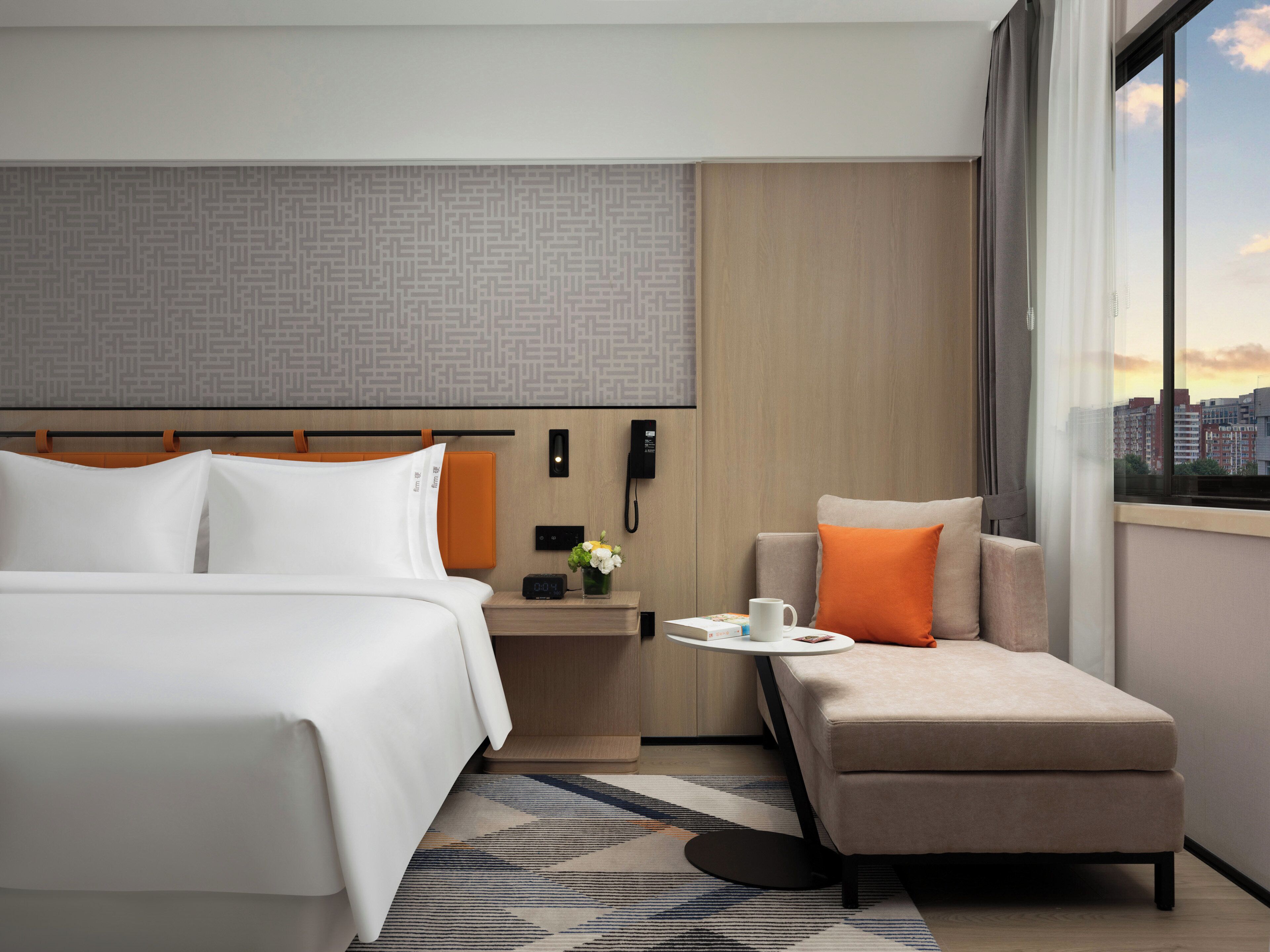 A hotel room with a bed, a sofa, a nightstand with a phone, a clock, and a window with a city view.