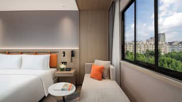A hotel room with a bed, a sofa, a small table, a nightstand, and a large window offering a city view.