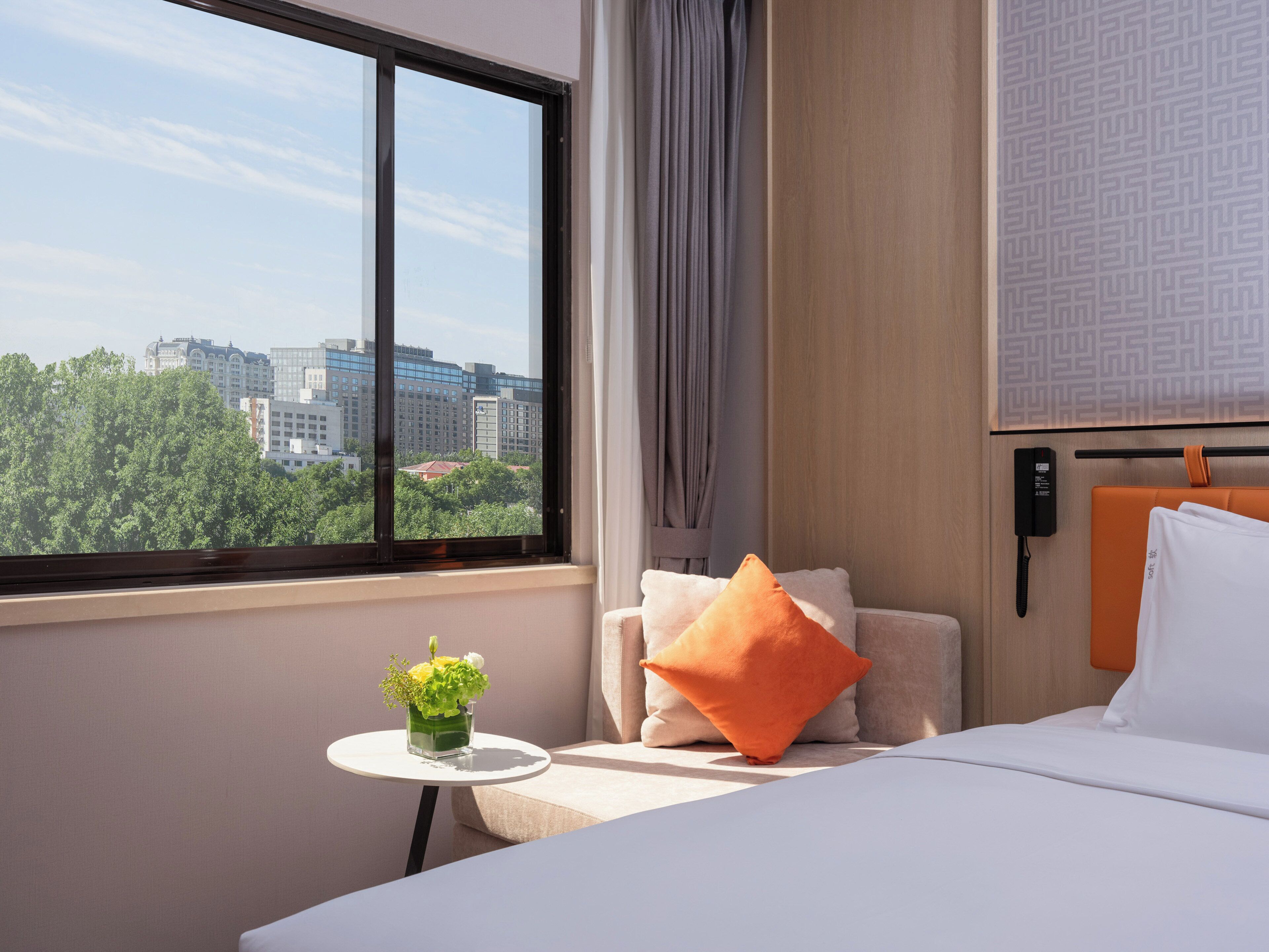 A hotel room with a bed, a small table with a vase of flowers, a chair with cushions, a window with a city view, and a wall-mounted phone.