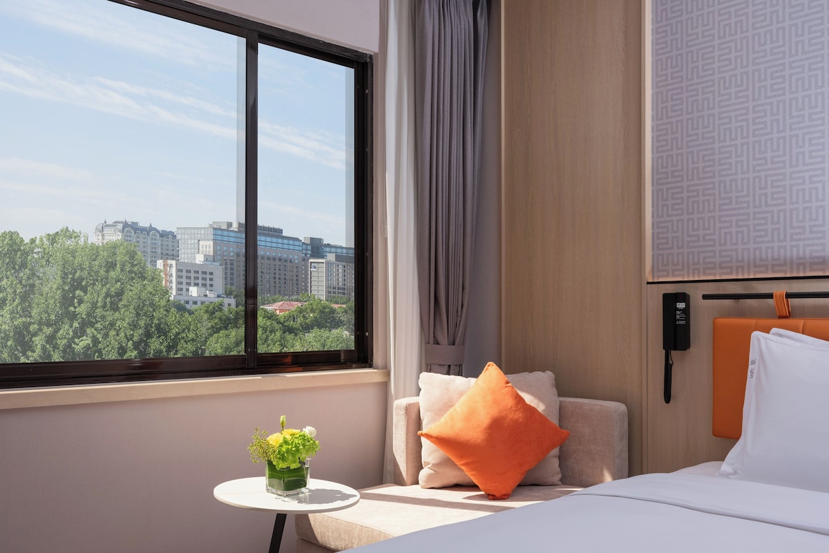 A hotel room with a bed, a small table with a vase of flowers, a chair with cushions, a window with a city view, and a wall-mounted phone.