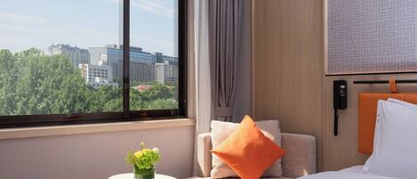 A hotel room with a bed, a small table with a vase of flowers, a chair with cushions, a window with a city view, and a wall-mounted phone.