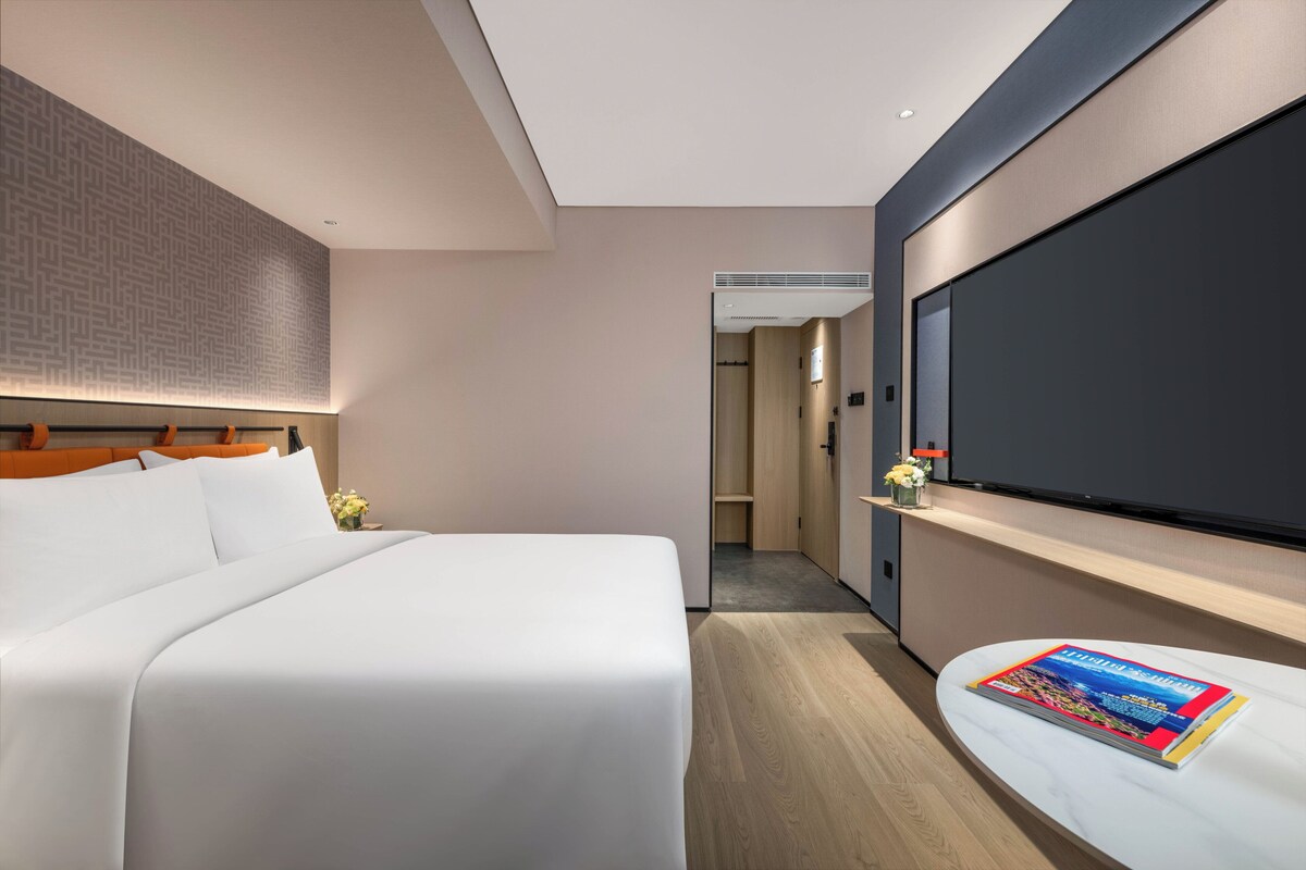 A modern hotel room with a large bed, a flat-screen TV mounted on the wall, and a magazine on the bedside table.
