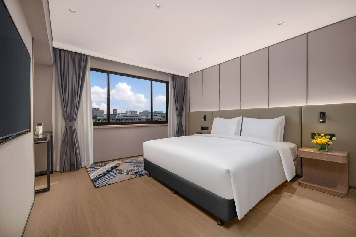 A modern hotel room with a large bed, a city view, and a small side table with a vase of flowers.