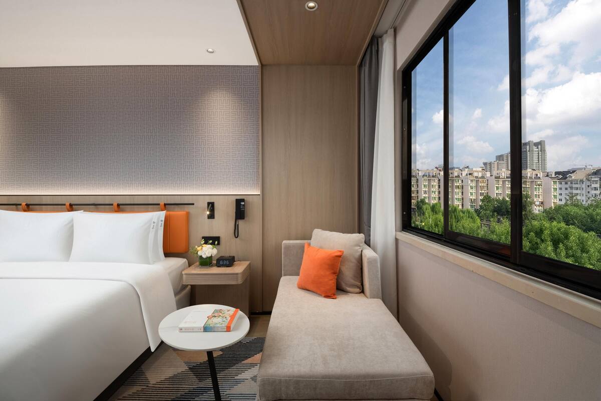 A hotel room with a bed, a sofa, a small table with a book, a nightstand with a phone, and a window with a city view.