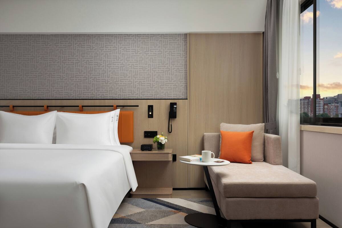 A hotel room with a bed, a sofa, a nightstand with a phone, a small table with a cup, and a window with a city view.