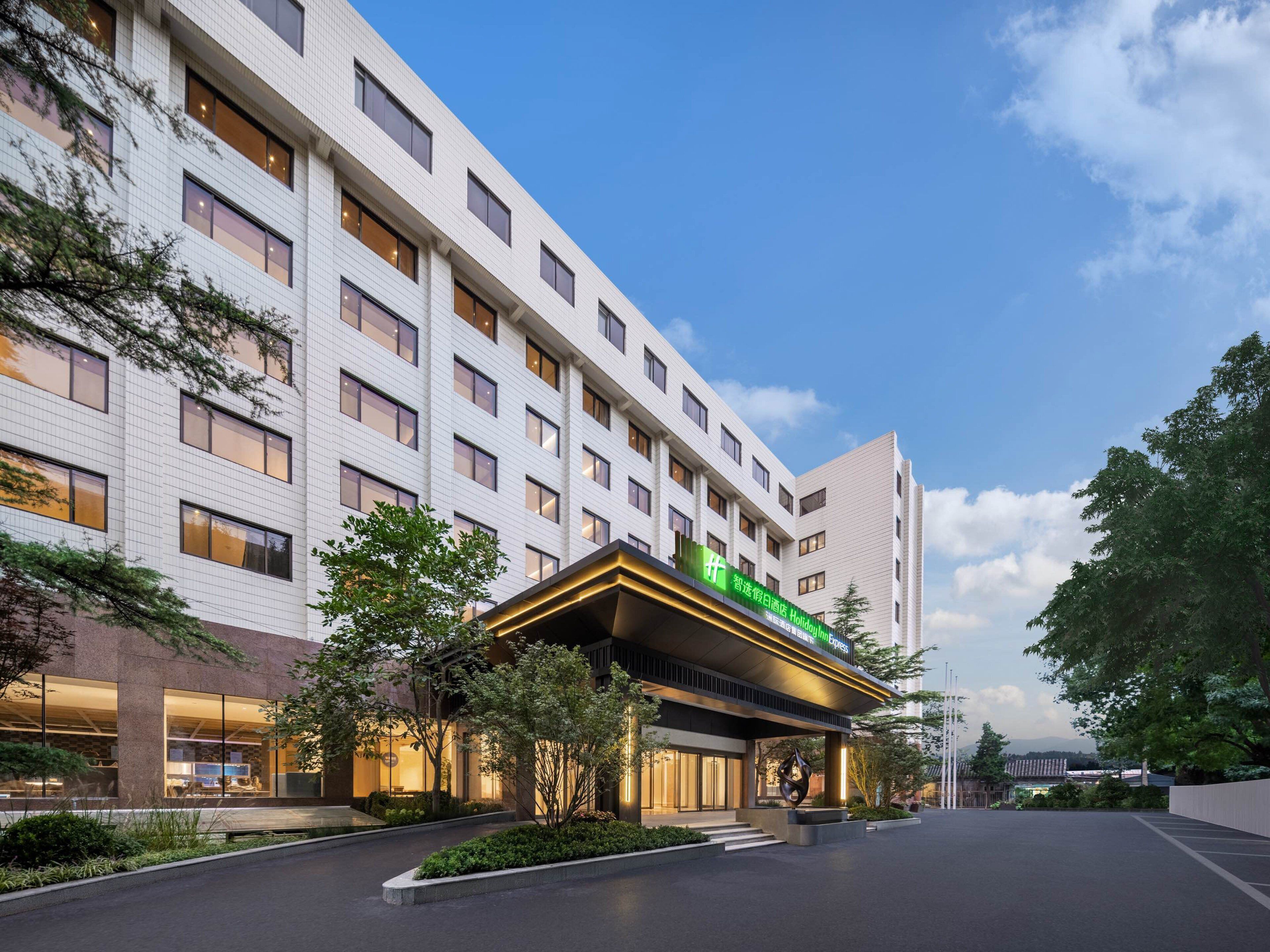 A modern hotel building with a prominent green sign, large windows, and a well-maintained garden area in front.