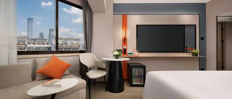 A modern hotel room with a large bed, a sofa, a small table, a chair, a TV, and a minibar.