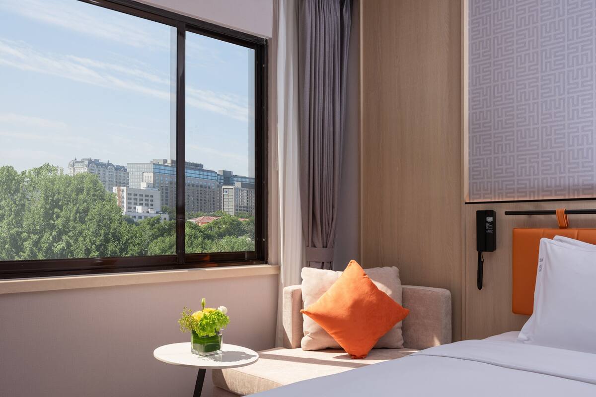 A hotel room with a bed, a small table with a vase of flowers, a chair with cushions, a window with a city view, and a wall-mounted phone.