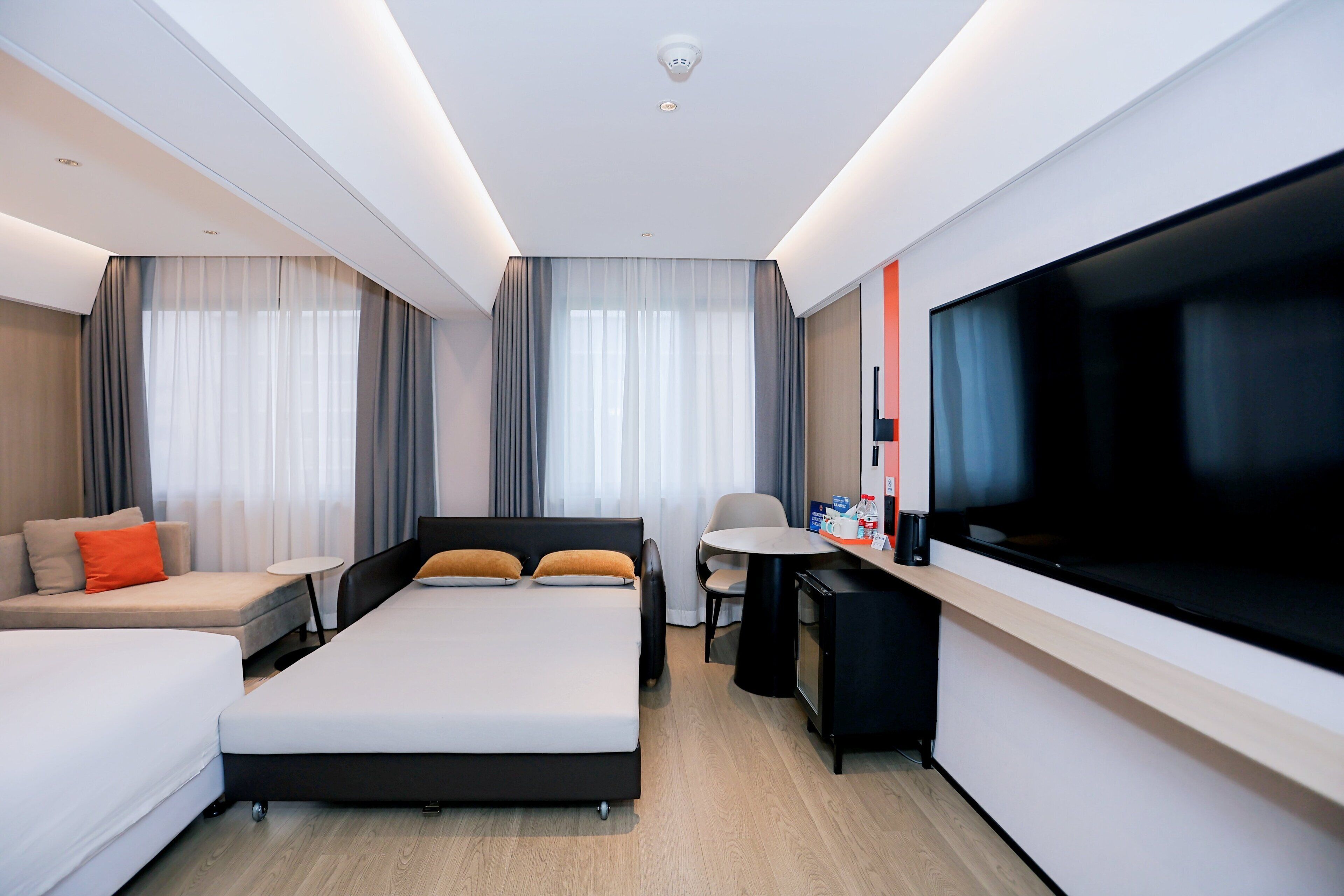 A modern hotel room with a large bed, a sofa, a desk with a flat-screen TV, and a small table.