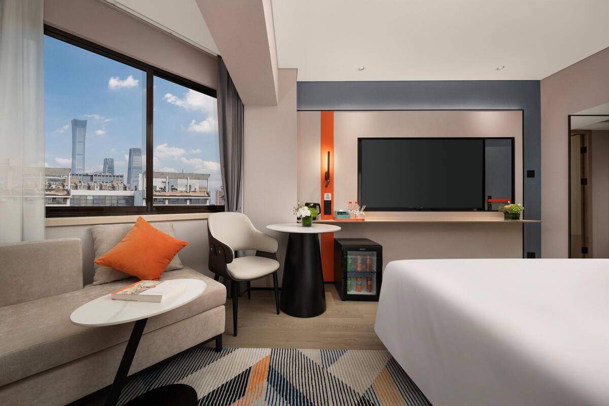 A modern hotel room with a large bed, a sofa, a small round table, a chair, a TV, a mini-fridge, and a window with a city view.