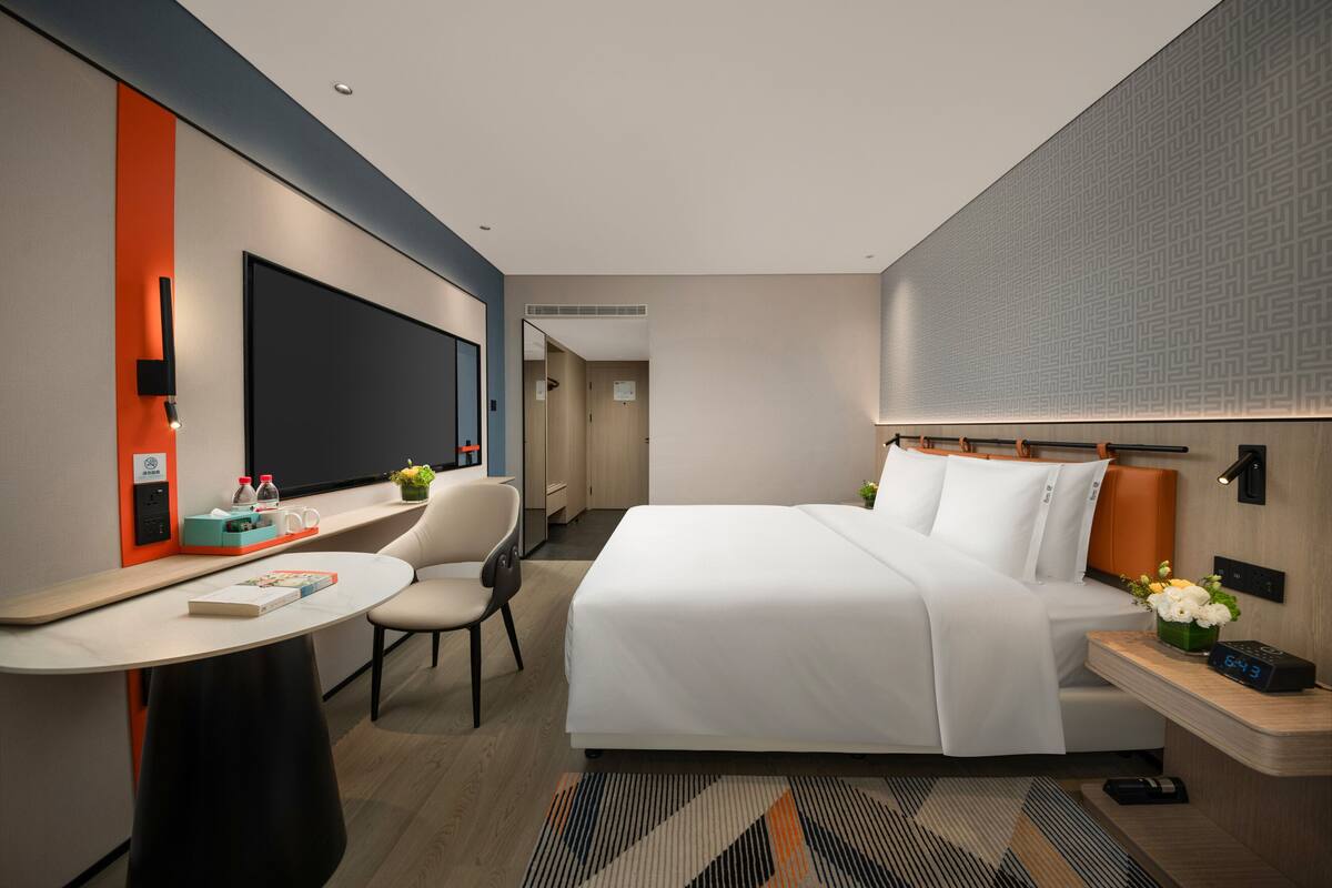 A modern hotel room with a bed, a desk, a chair, a TV, and a nightstand with a clock.