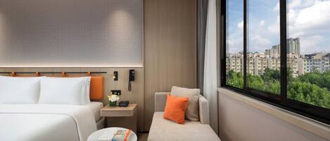 Ein Hotelzimmer mit einem Bett, einer Couch, einem kleinen Tisch mit einem Buch, einem Nachttisch mit einem Telefon und einem Fenster mit Blick auf die Stadt.