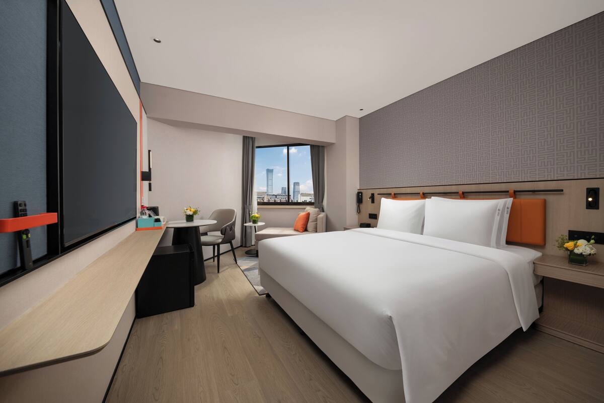 A modern hotel room with a large bed, a desk with a chair, a small table with a vase of flowers, and a view of the cityscape through a large window.