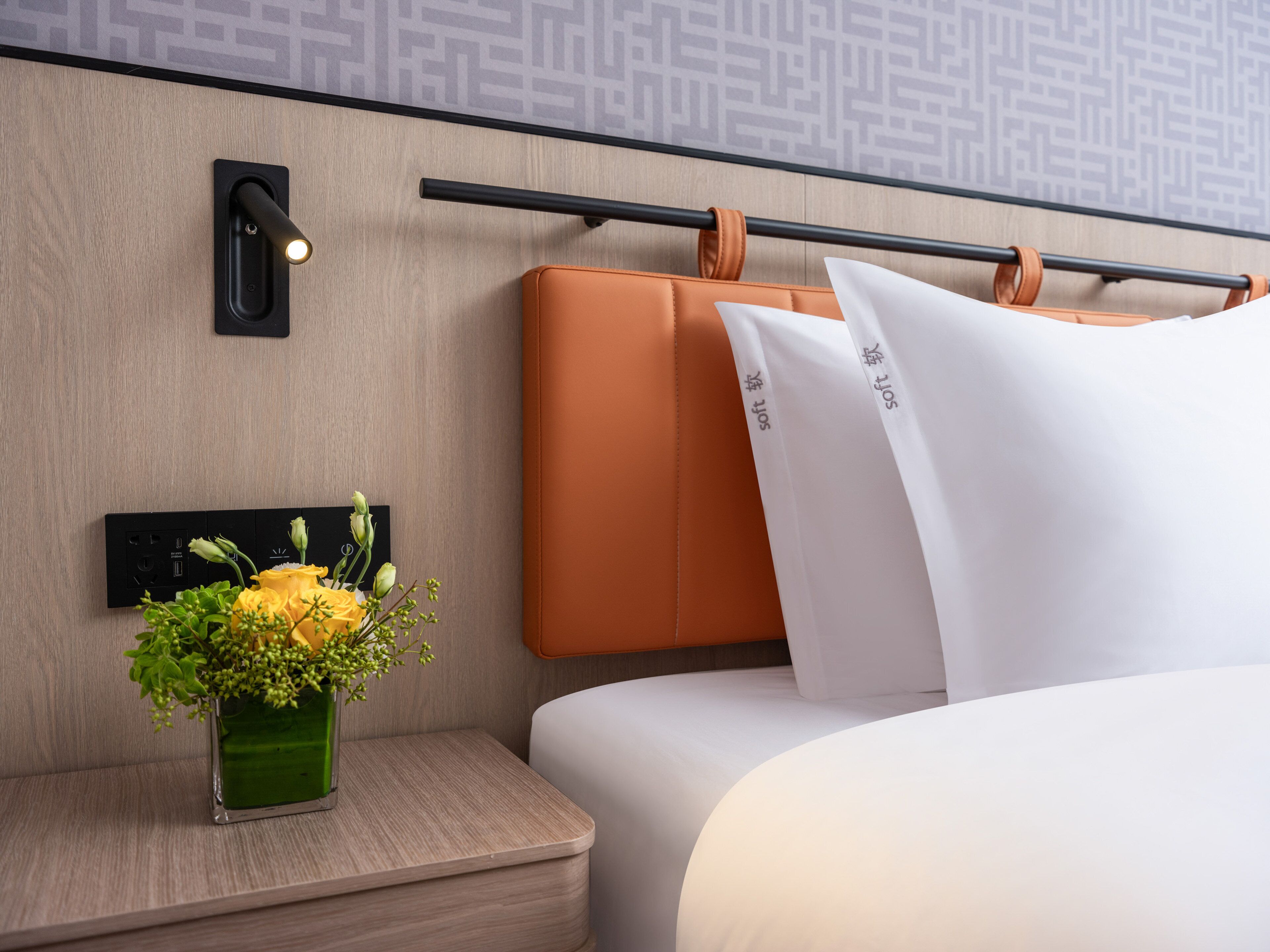 A modern hotel room with a wooden headboard, a bedside table with a vase of yellow flowers, and a black wall-mounted light.