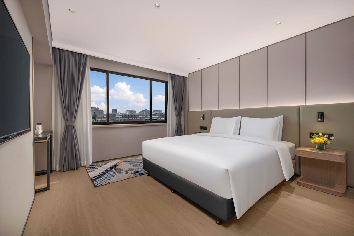 A modern hotel room with a large bed, a view of the cityscape, and a small table with a vase of flowers.