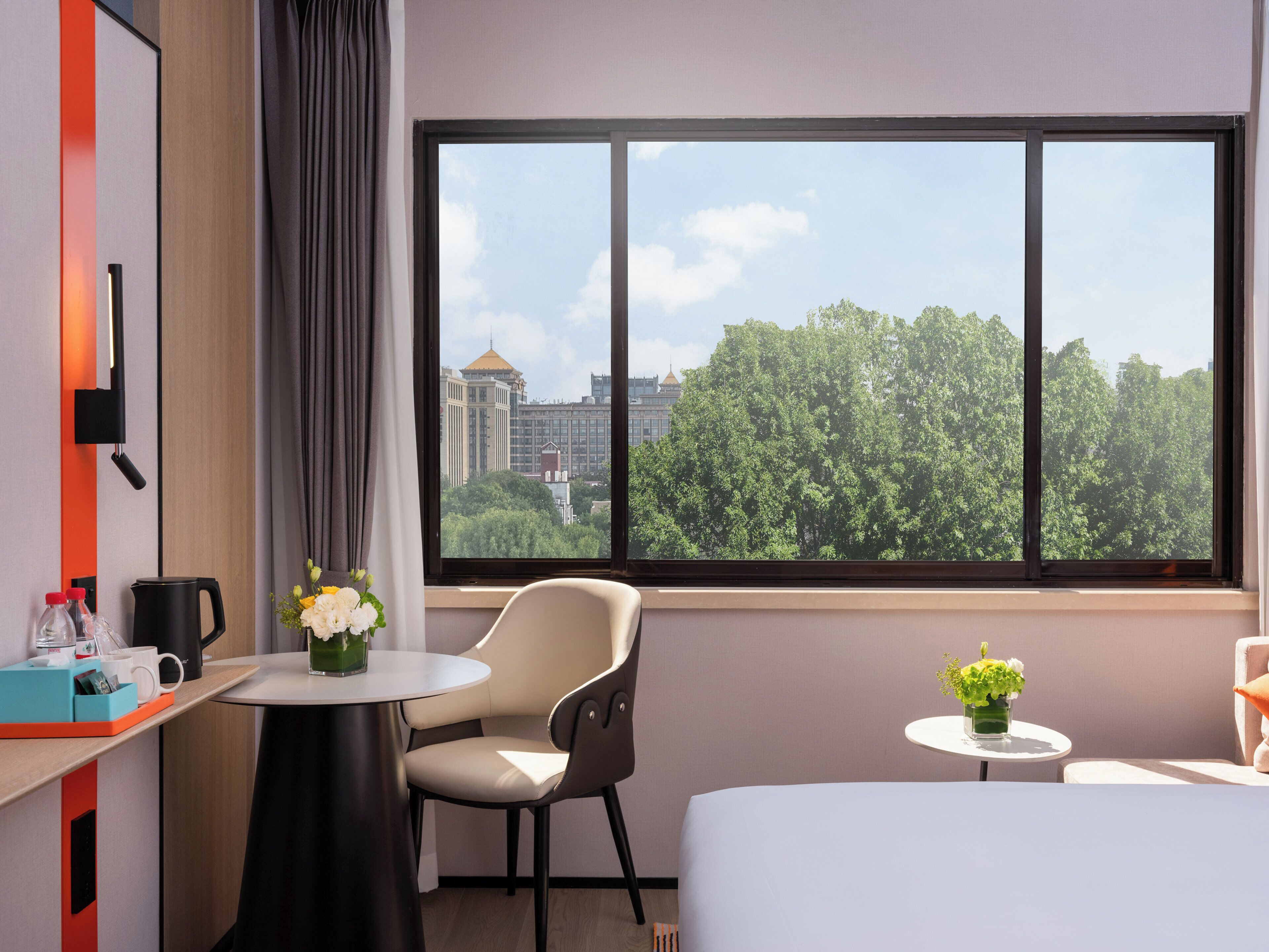 A hotel room with a large window, a desk with a chair, a bed, and a small table with a vase of flowers.