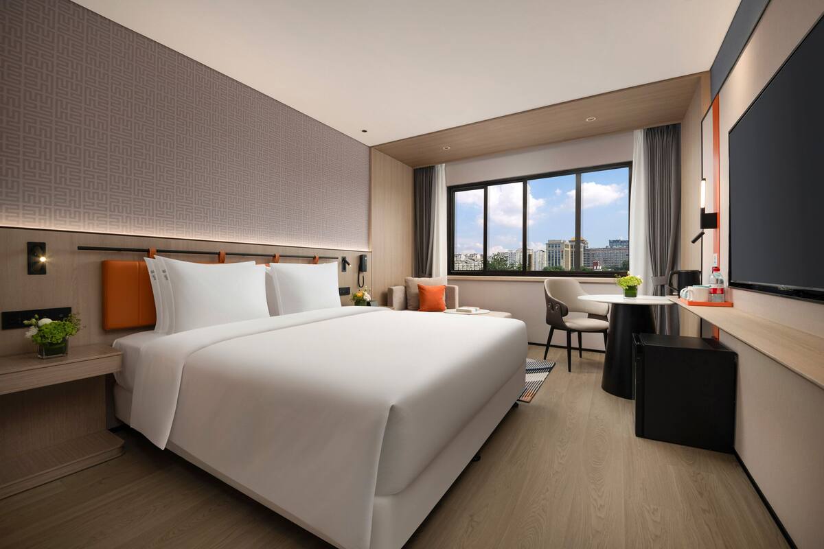 A modern hotel room with a large bed, a desk, and a view of the city.