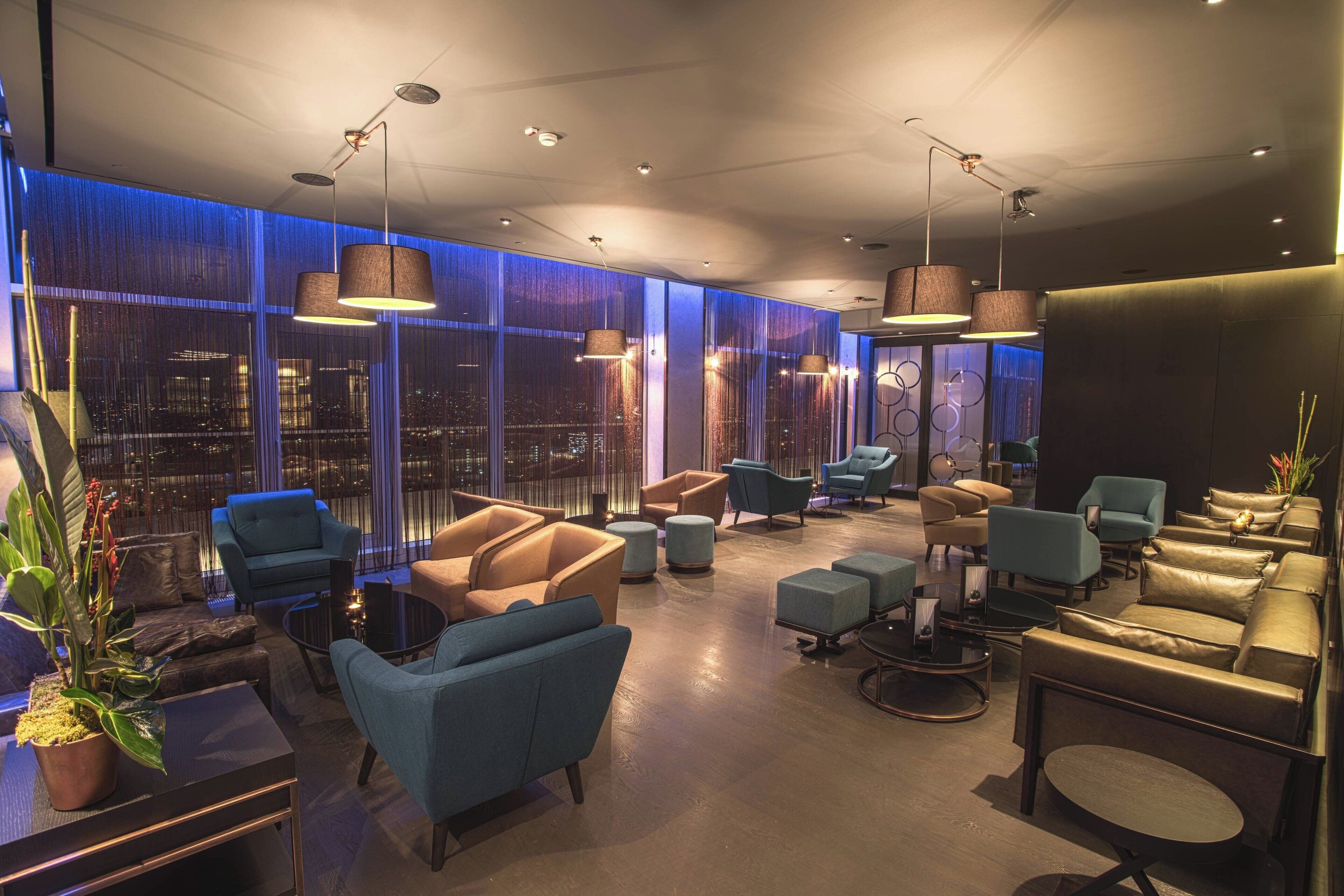 A modern lounge area with multiple seating options, including sofas and armchairs, arranged around a central coffee table. Large windows provide a view of the cityscape.