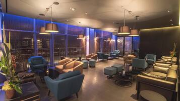 A modern lounge area with multiple seating options, including sofas and armchairs, arranged around a central coffee table. Large windows provide a view of the cityscape.