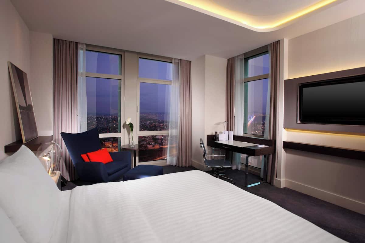 A hotel room with a large bed, a desk, a chair, a TV, and a view of the city at night.