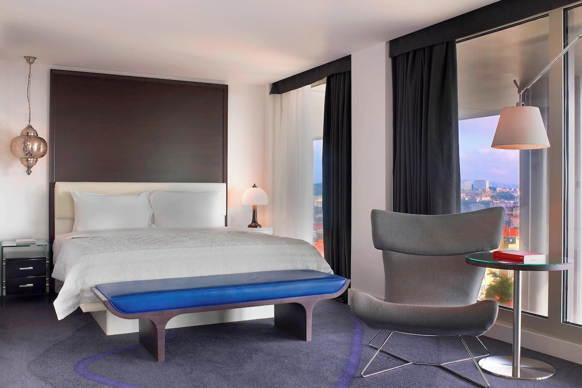 A modern hotel room with a large bed, a bench, a chair, a small table, a lamp, and a view of the city through large windows.