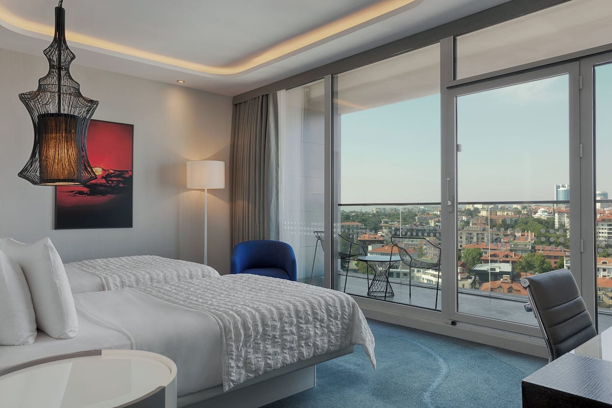 A modern hotel room with a large bed, a desk, and a balcony with a city view.