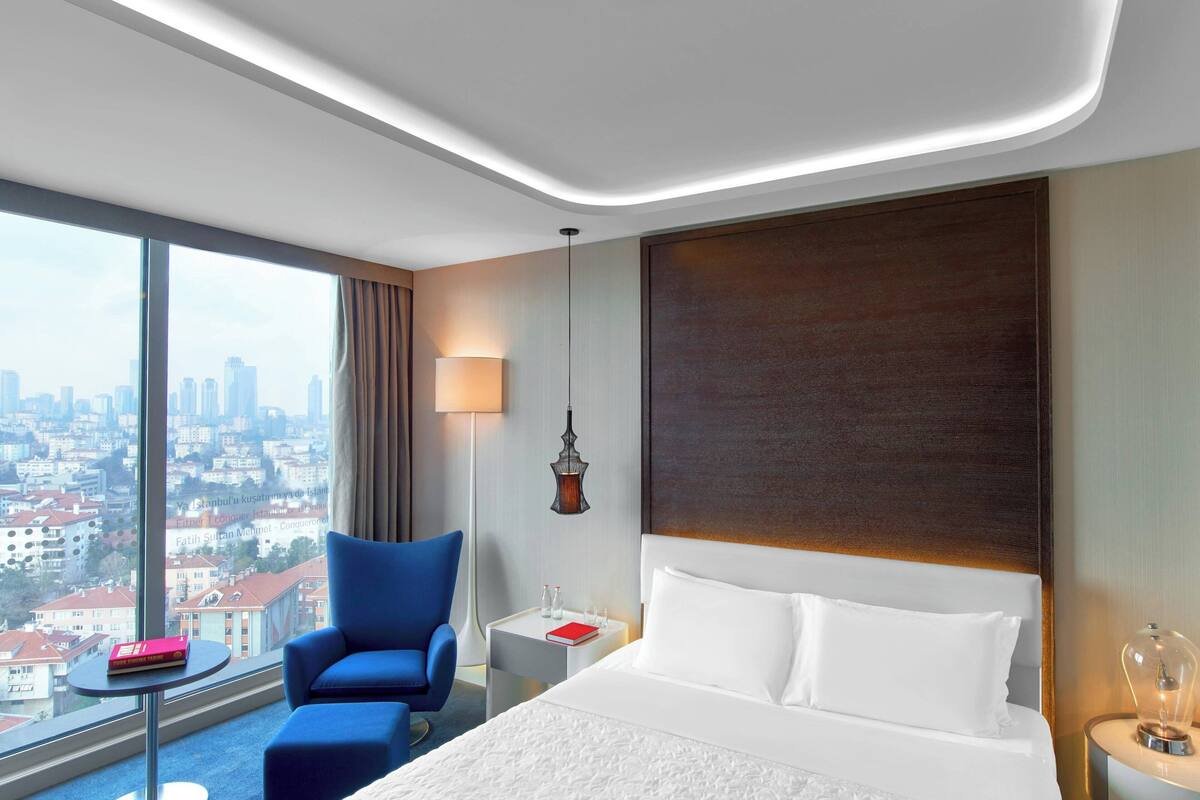 A hotel room with a large bed, a blue armchair, a small table with a red book, a nightstand with a lamp, and a view of a cityscape through large windows.