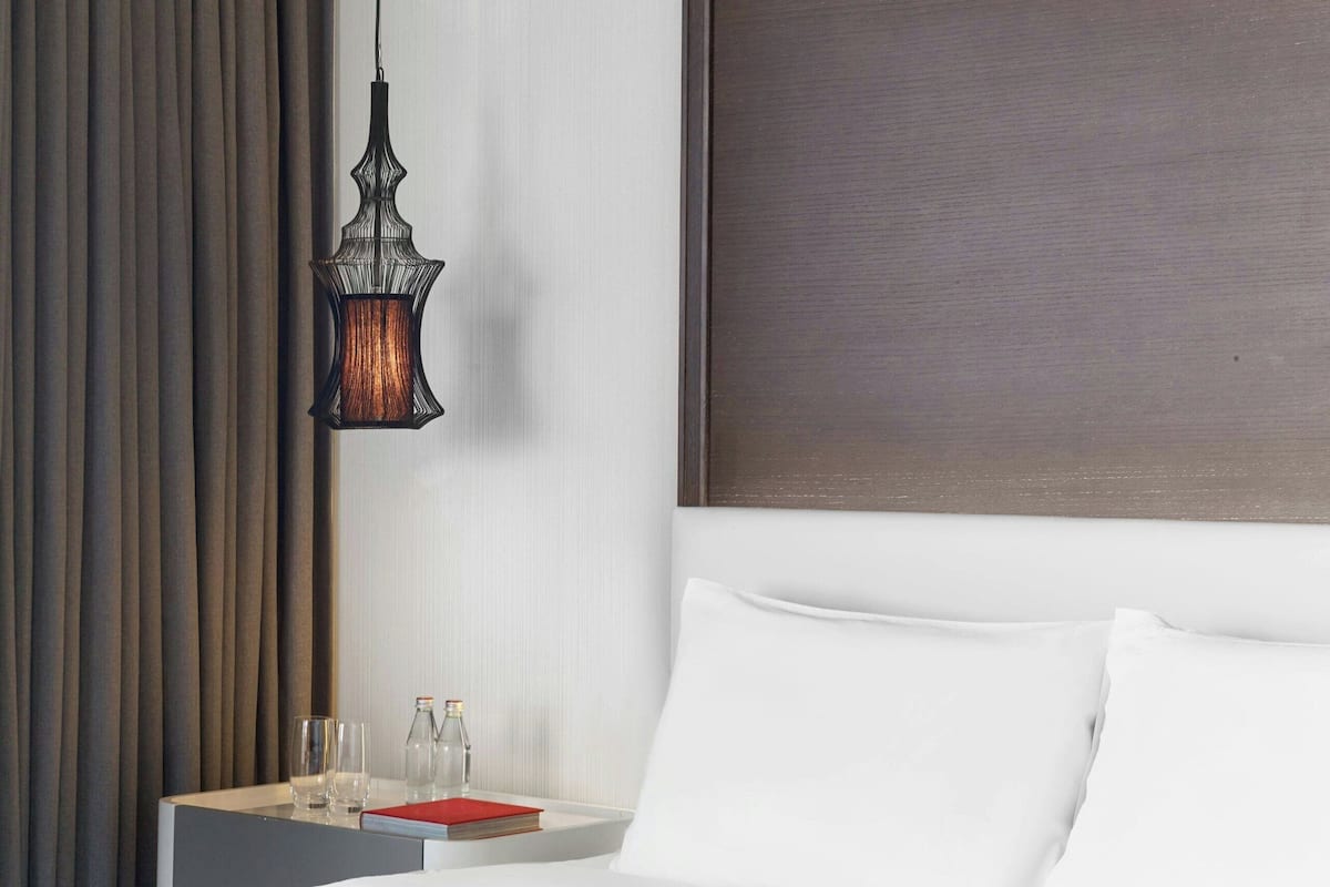 A hotel room with a large bed, a blue armchair, a nightstand with glasses and a bottle, and a hanging lamp.