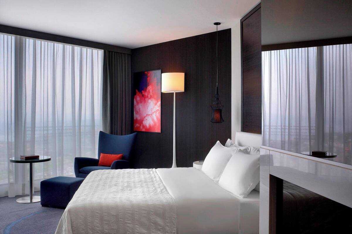A modern hotel room with a large bed, a seating area with a chair and ottoman, a standing lamp, and a large window with curtains.