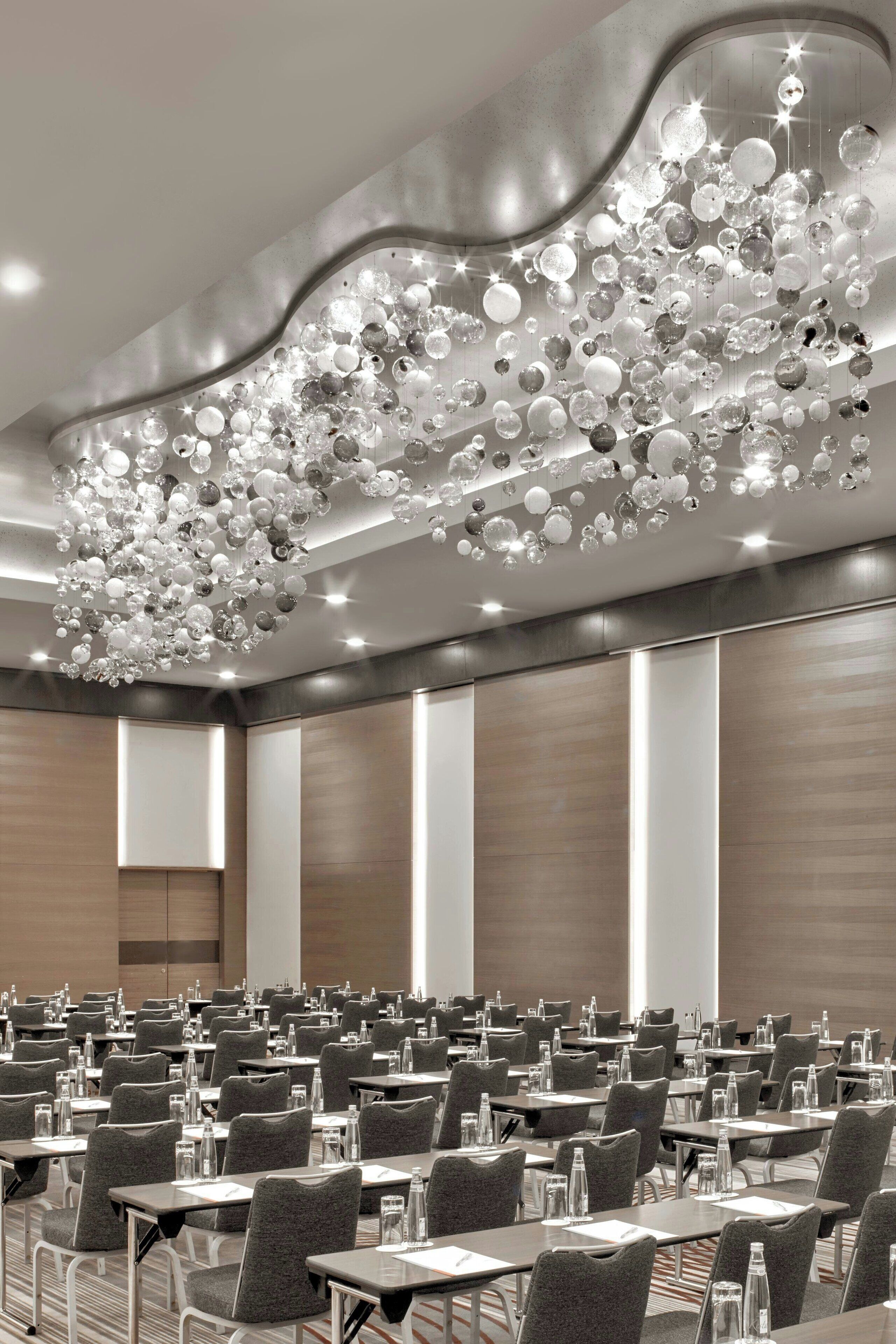 A conference room with rows of tables and chairs, a modern chandelier, and wooden panel walls.