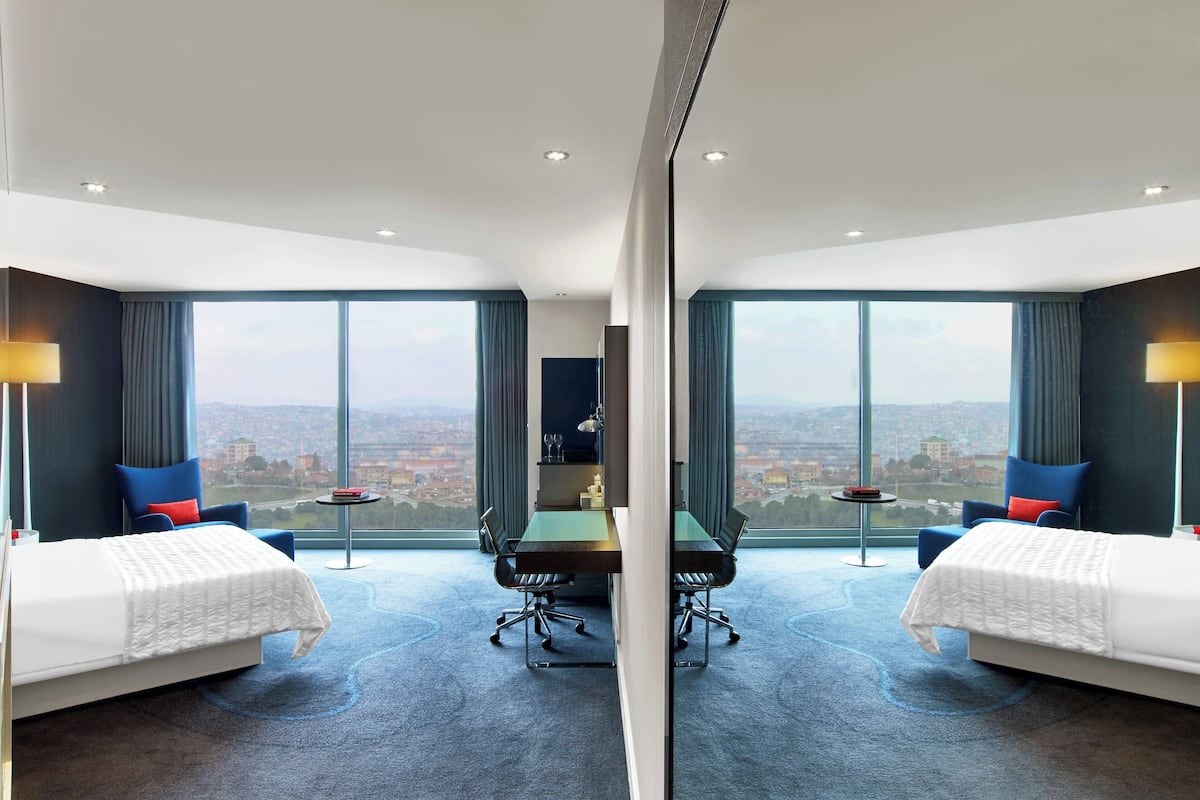 A hotel room with two beds, a dining table, and a view of a cityscape.