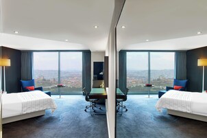 Habitación de hotel con dos camas, una mesa de comedor y vista a un paisaje urbano.