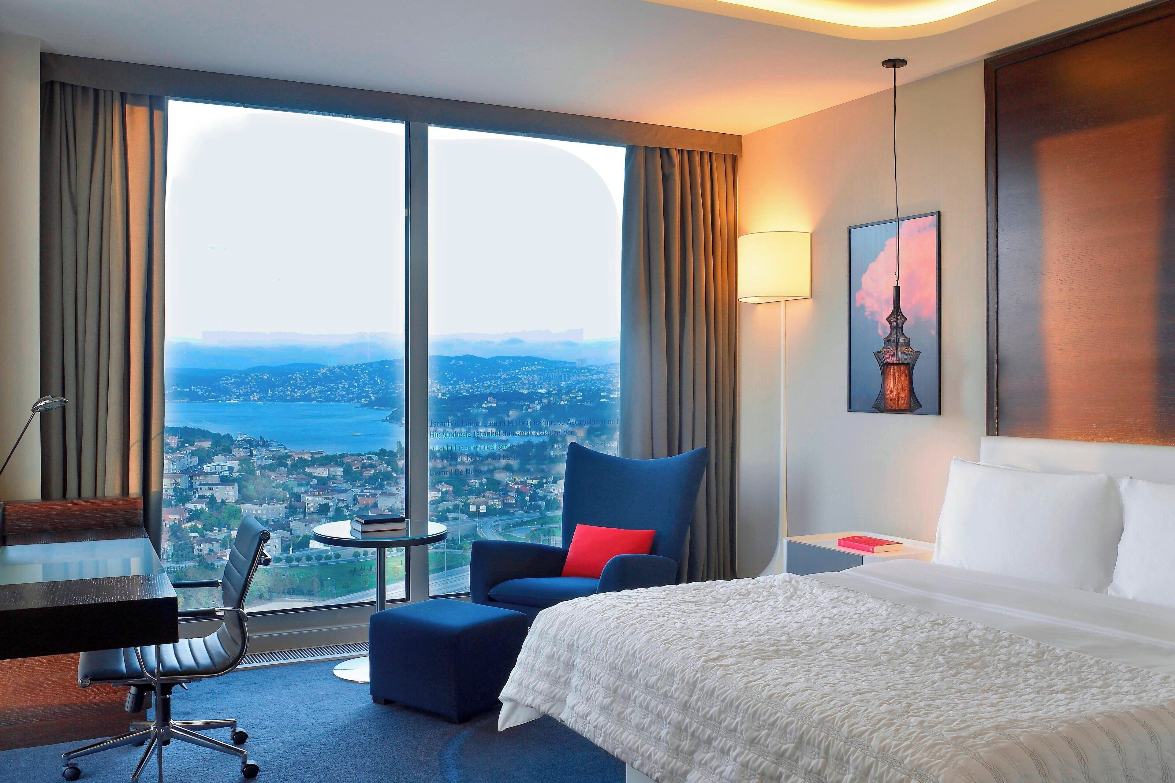 A hotel room with a large bed, a desk, a chair, and a view of a cityscape through large windows.