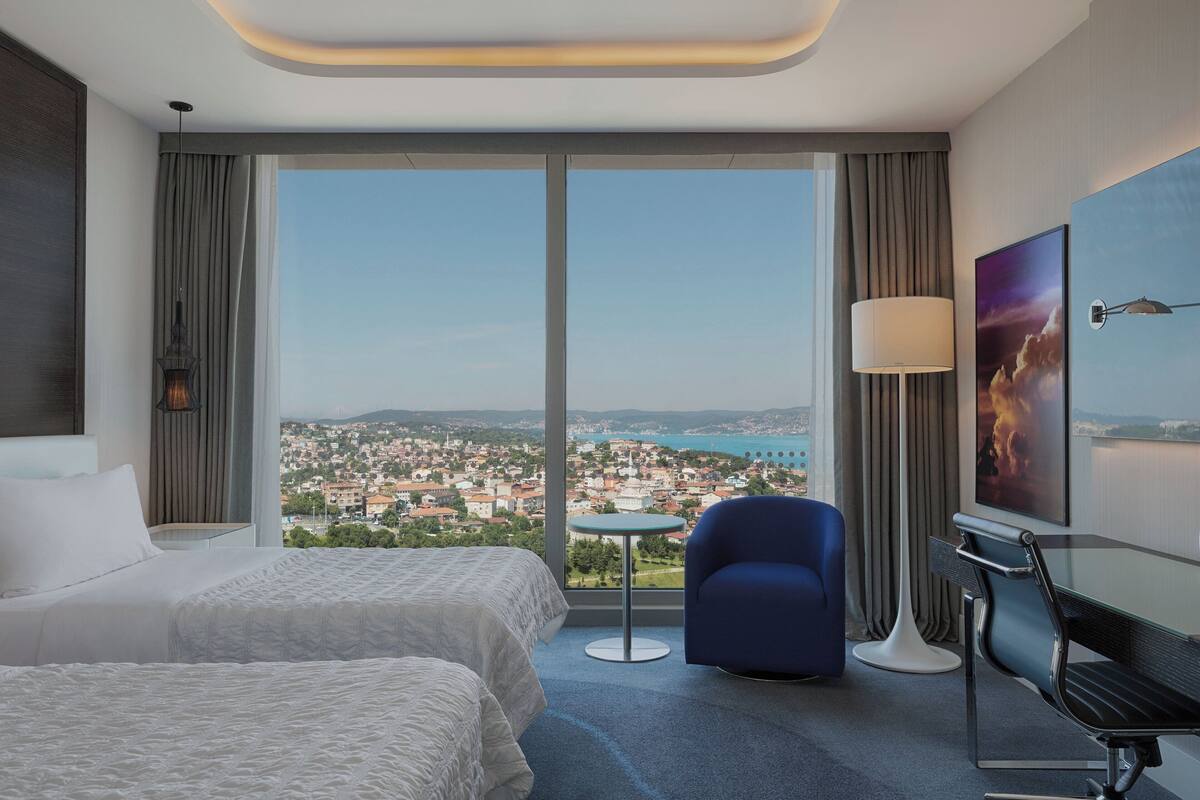 A hotel room with two beds, a view of a cityscape, a round table, a blue chair, a floor lamp, and a TV.