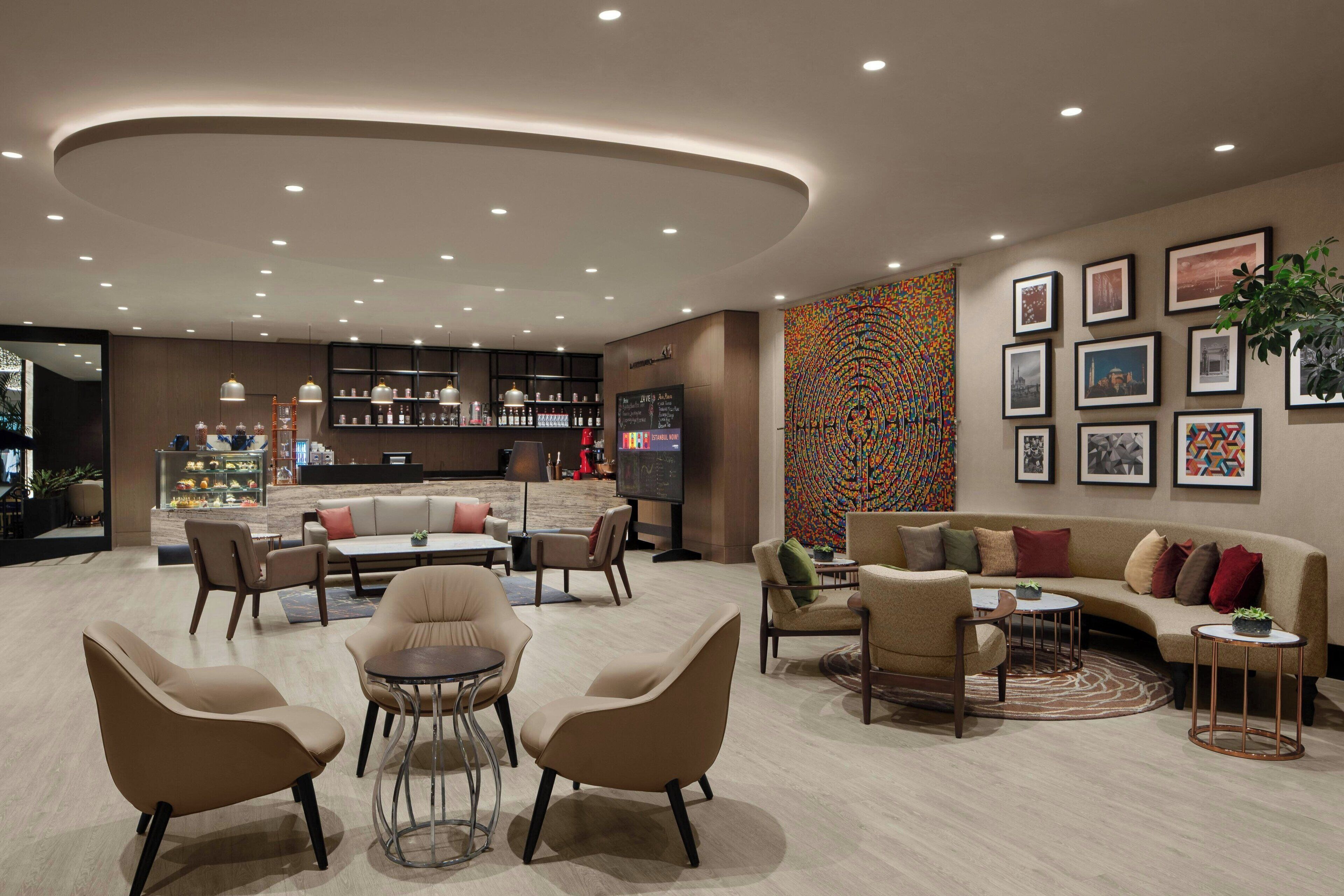 Un moderno vestíbulo de hotel con techo curvo, una zona de estar central con sofás y sillones, un área de bar con estanterías y un televisor, y una llamativa obra de arte abstracta en la pared.