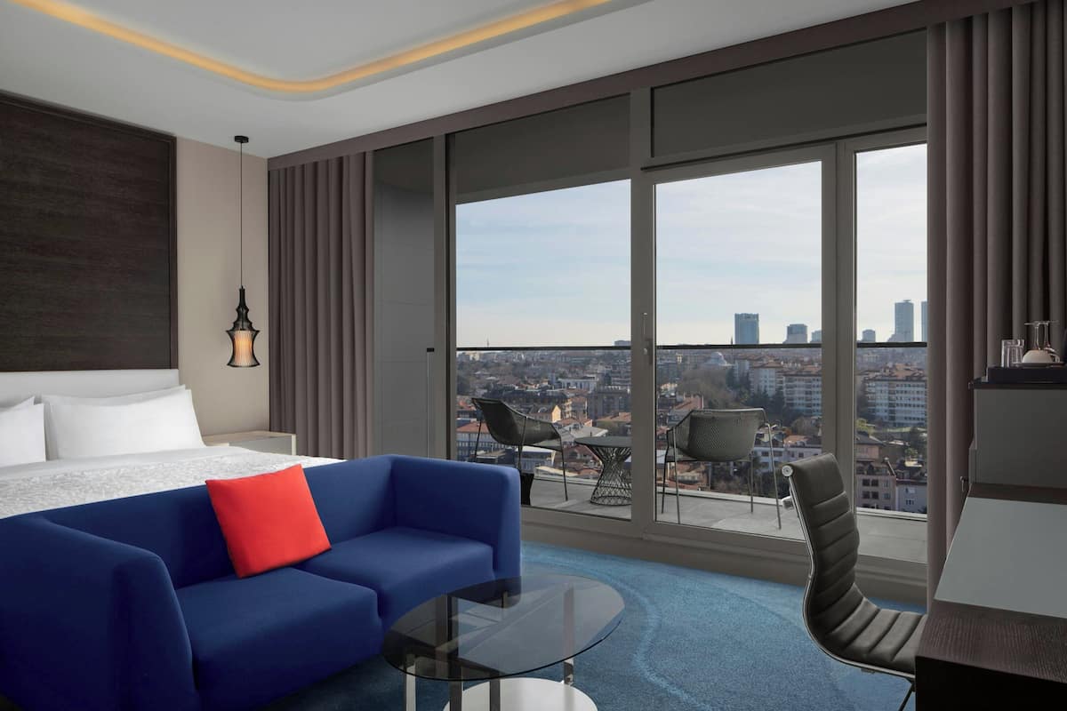 A modern hotel room with a large bed, a blue sofa, a glass coffee table, and a view of the city through large windows.