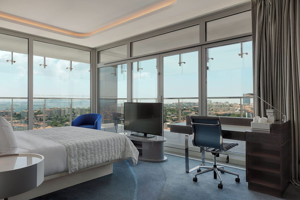 A modern hotel room with a large bed, a desk, a chair, a TV, and a view of the city.